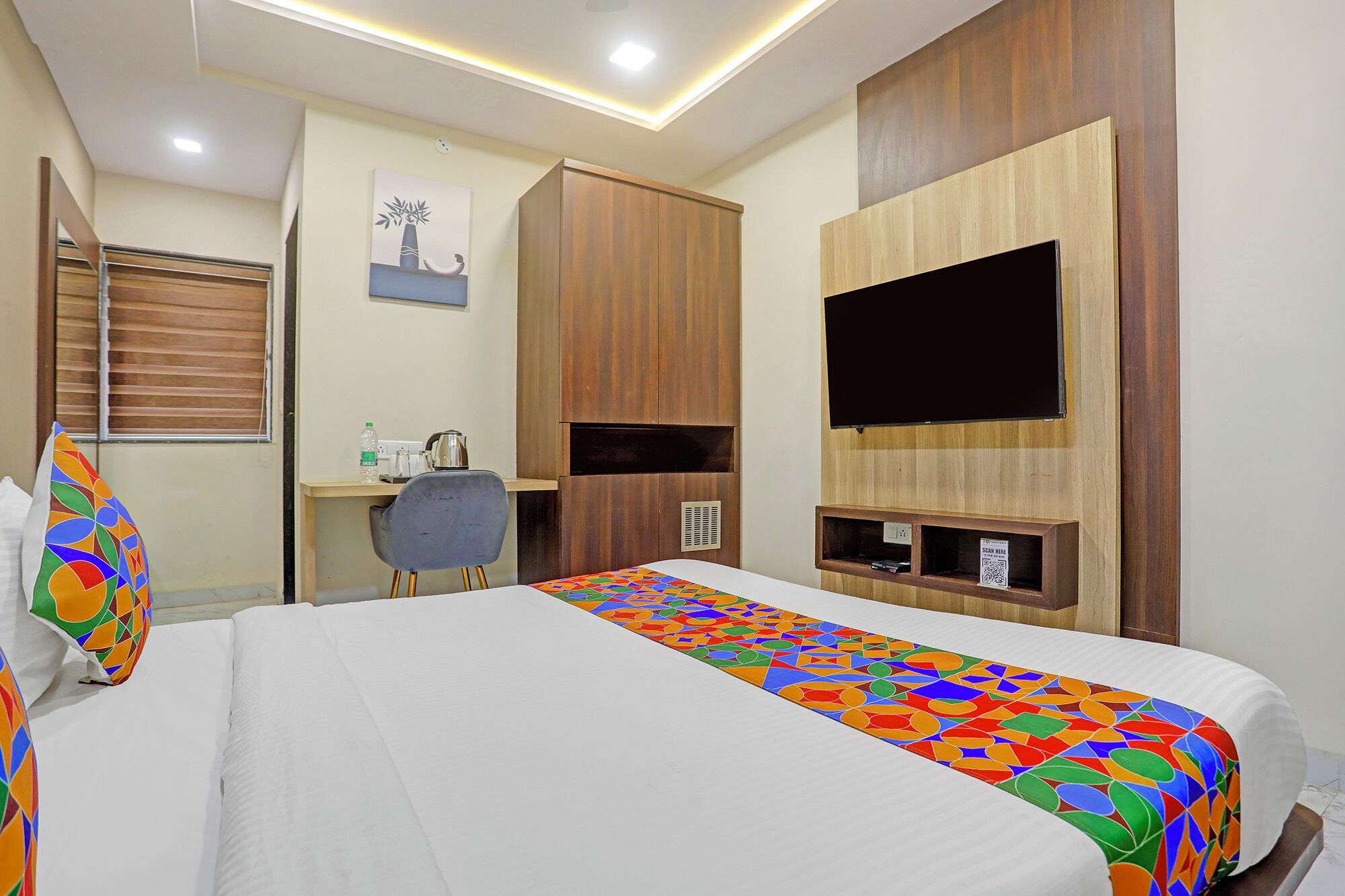 Habitación de hotel con cama, escritorio, silla, televisor y ventana con persianas.