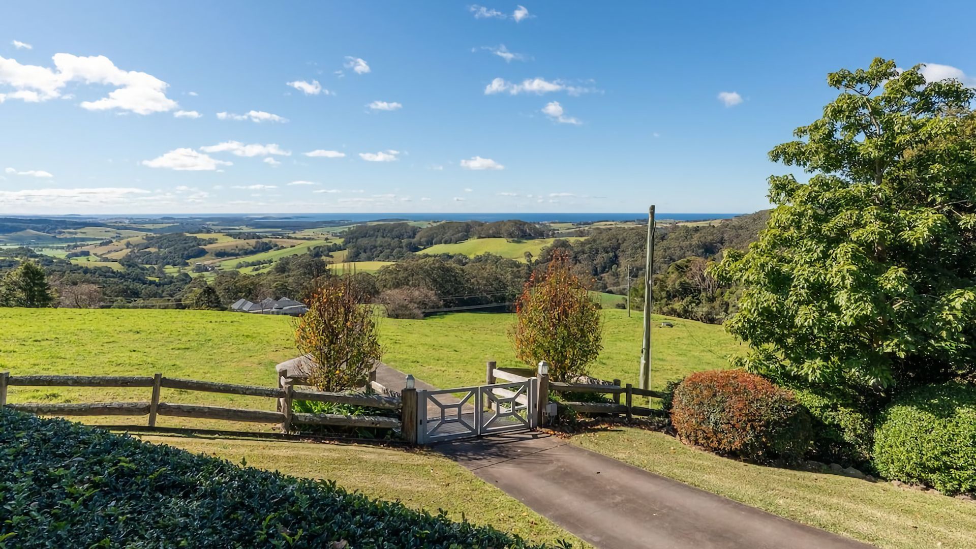 A rural landscape with a paved path leading to a gate, surrounded by greenery and a view of rolling hills.