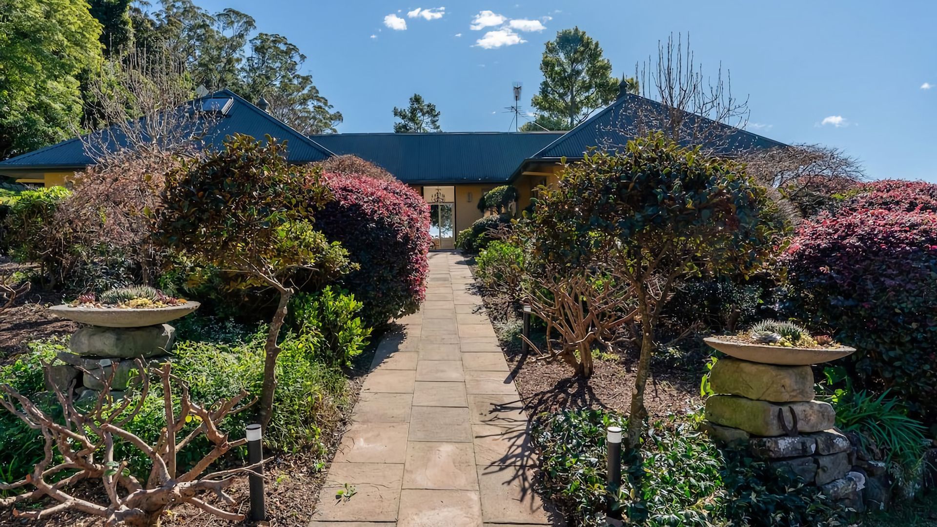 A pathway leading to a building with a dark roof, surrounded by various shrubs and plants.