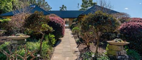 A pathway leading to a building with a dark roof, surrounded by various shrubs and plants.