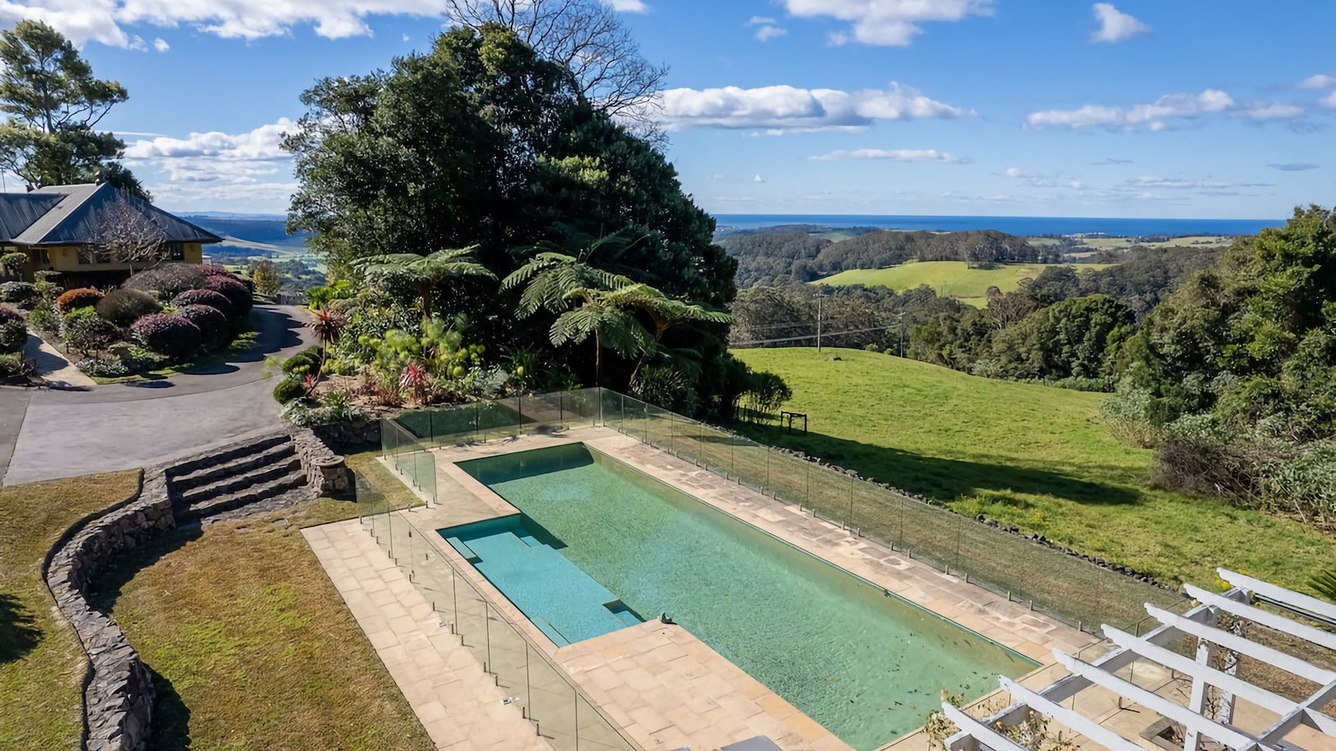 A swimming pool with a stone deck and glass railing, surrounded by lush greenery and a view of the ocean.