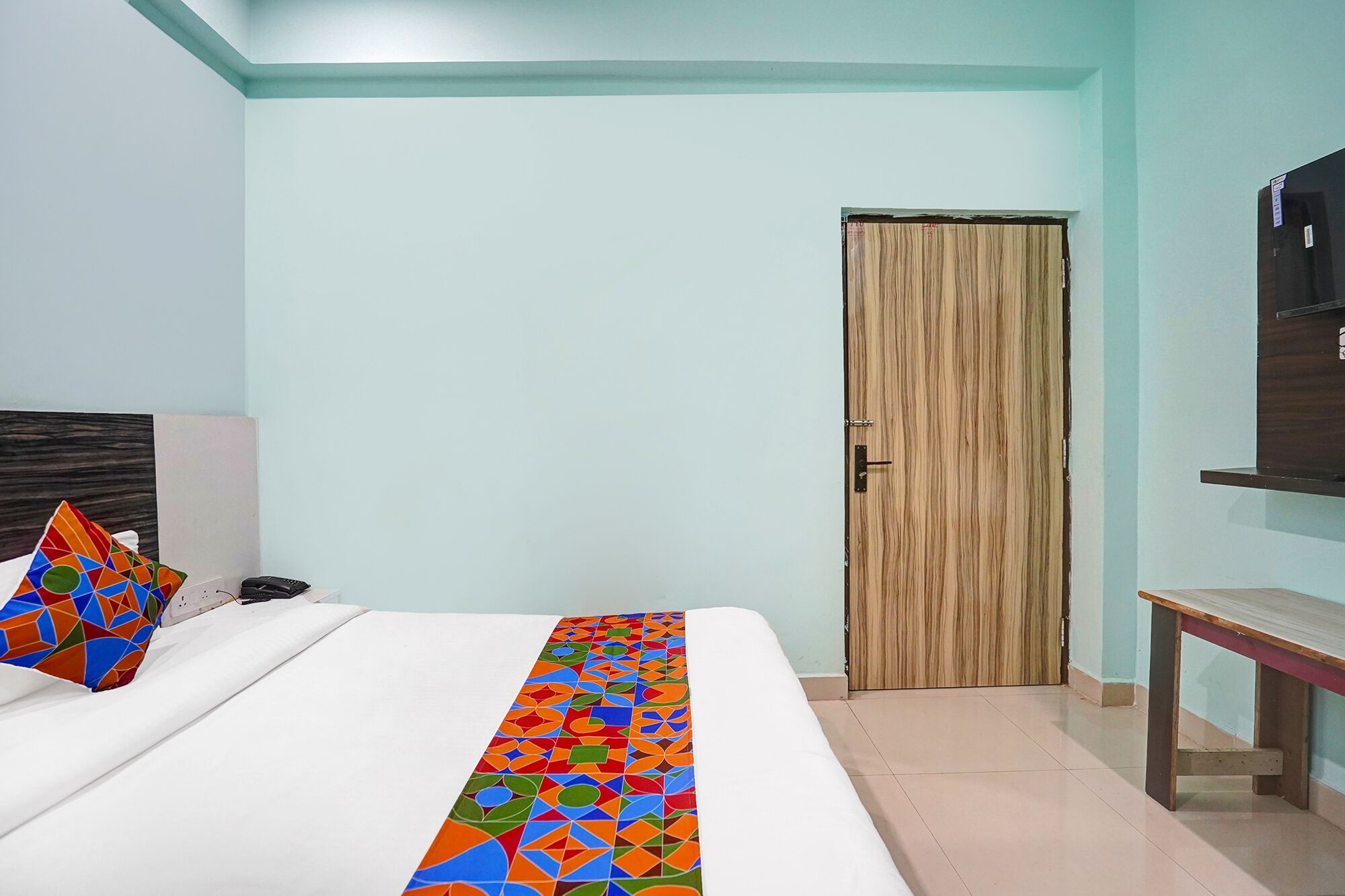 A hotel room with a bed, a wooden door, a bedside table, and a wall-mounted TV.