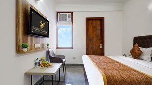 A hotel room with a bed, a desk with fruit, a chair, a TV on the wall, and a window with an air conditioner. - Fabhotel La Corporate Suite (Noida)