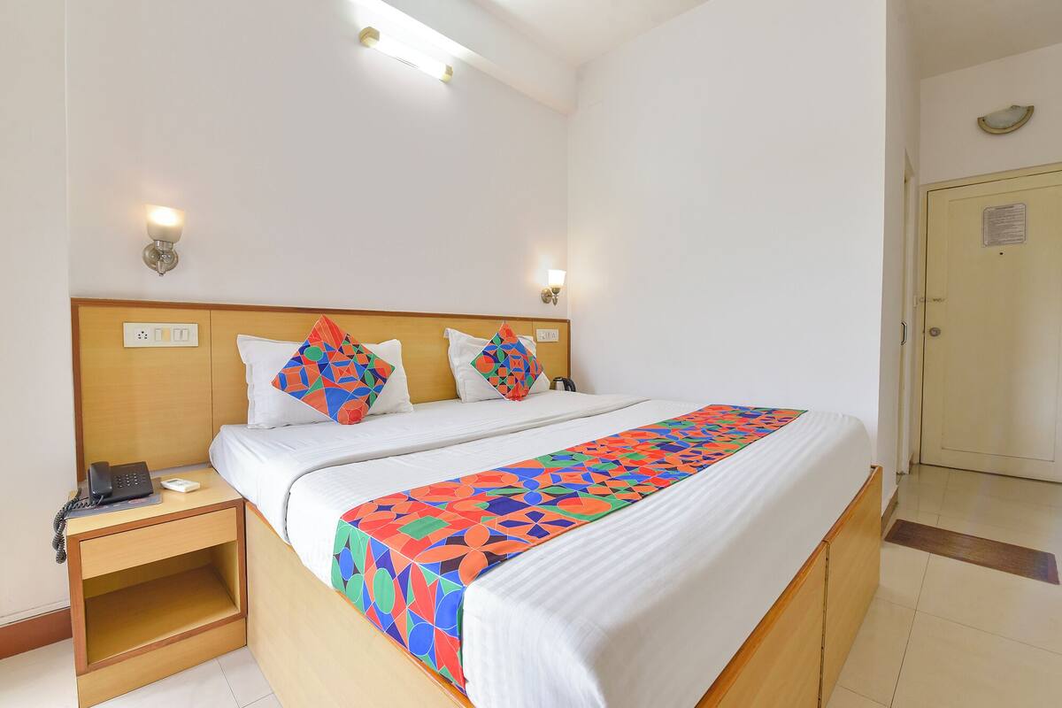 Une chambre d’hôtel avec une tête de lit en bois, du linge de lit blanc, une couvre-lit aux motifs géométriques colorés, une table de chevet avec un téléphone et une porte avec une pancarte.