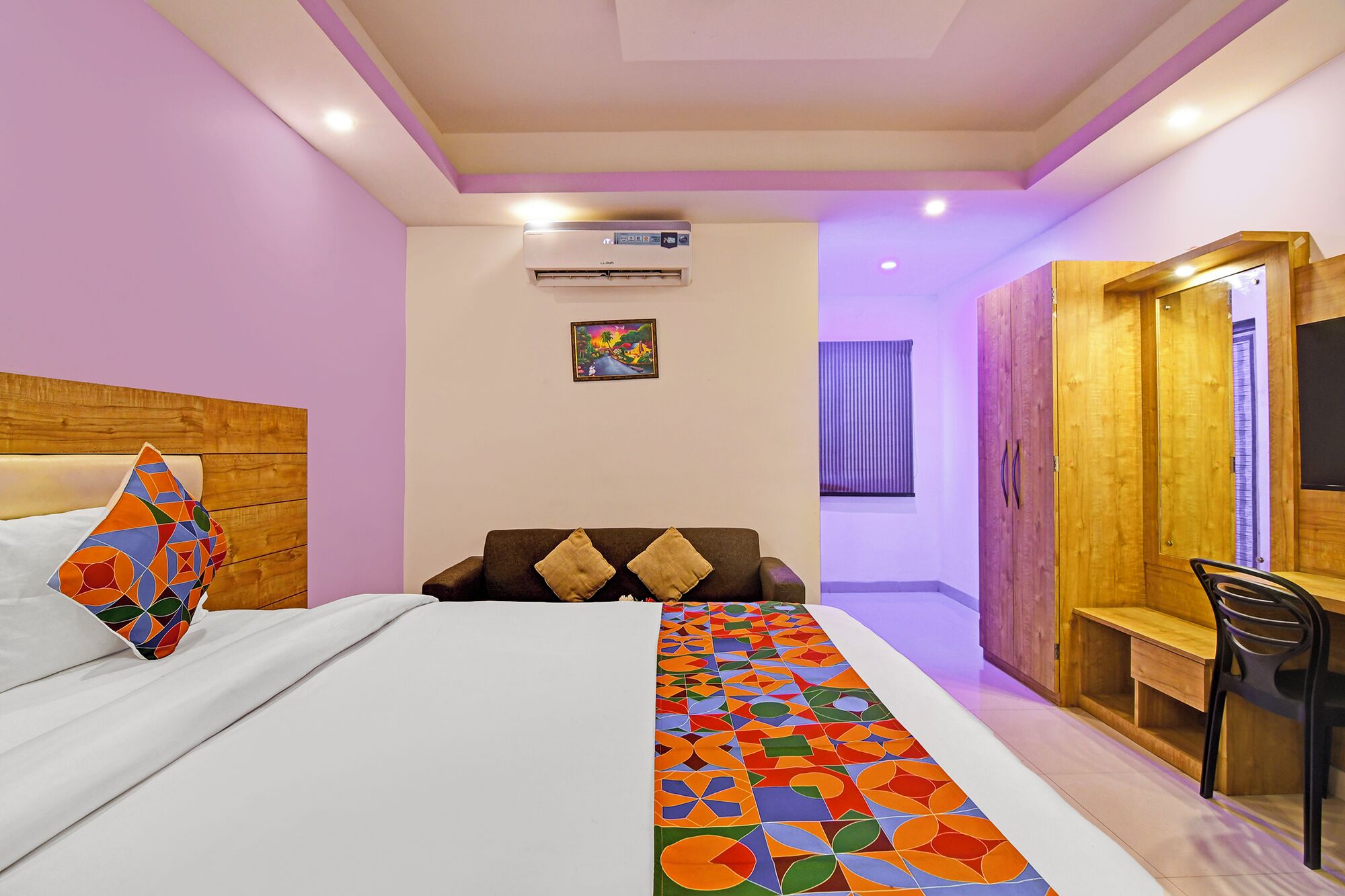 A hotel room with a bed, a wooden headboard, a colorful runner, a desk with a chair, a wardrobe, and an air conditioning unit.