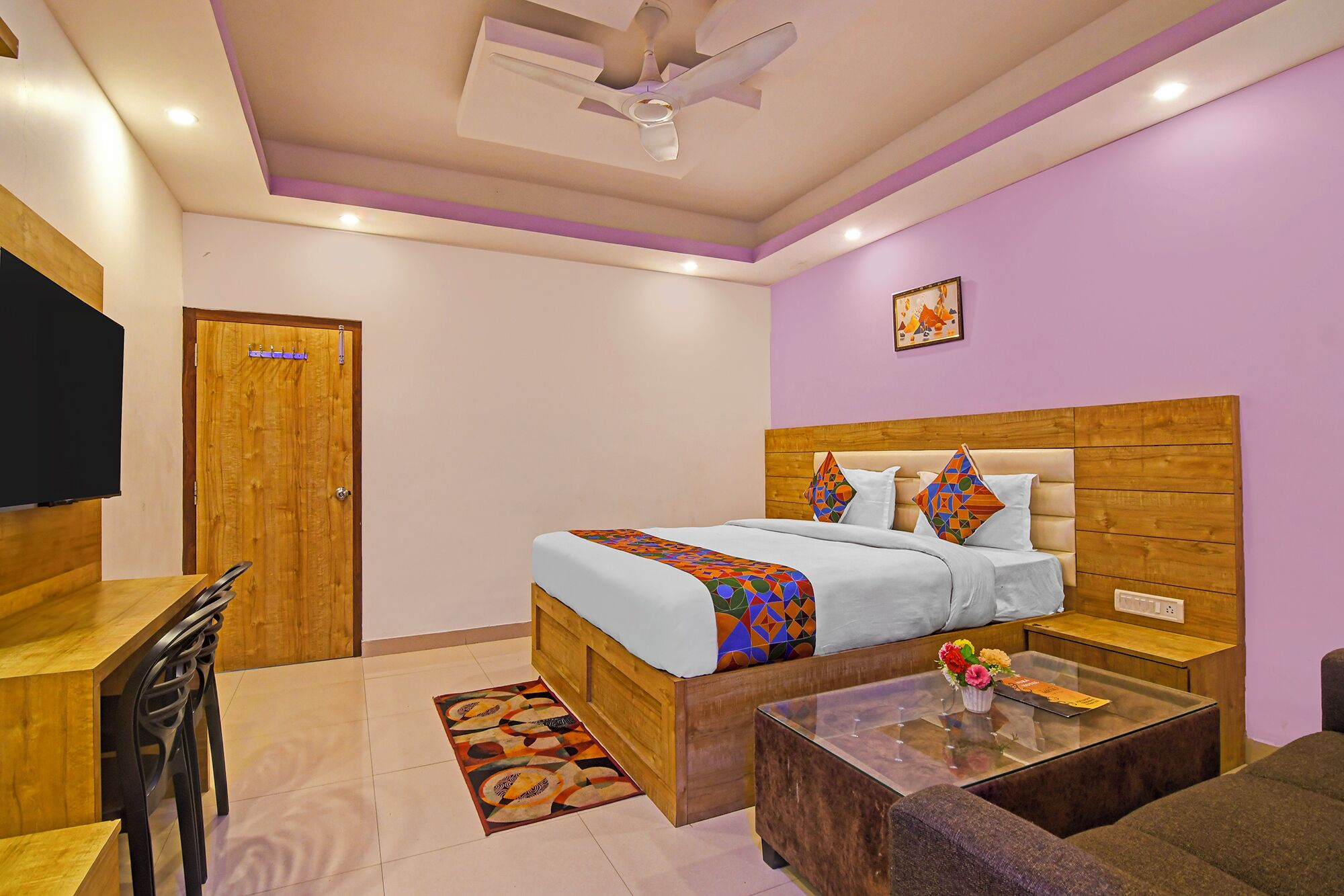 A bedroom with a wooden bed, a glass coffee table, a sofa, a TV, and a ceiling fan.