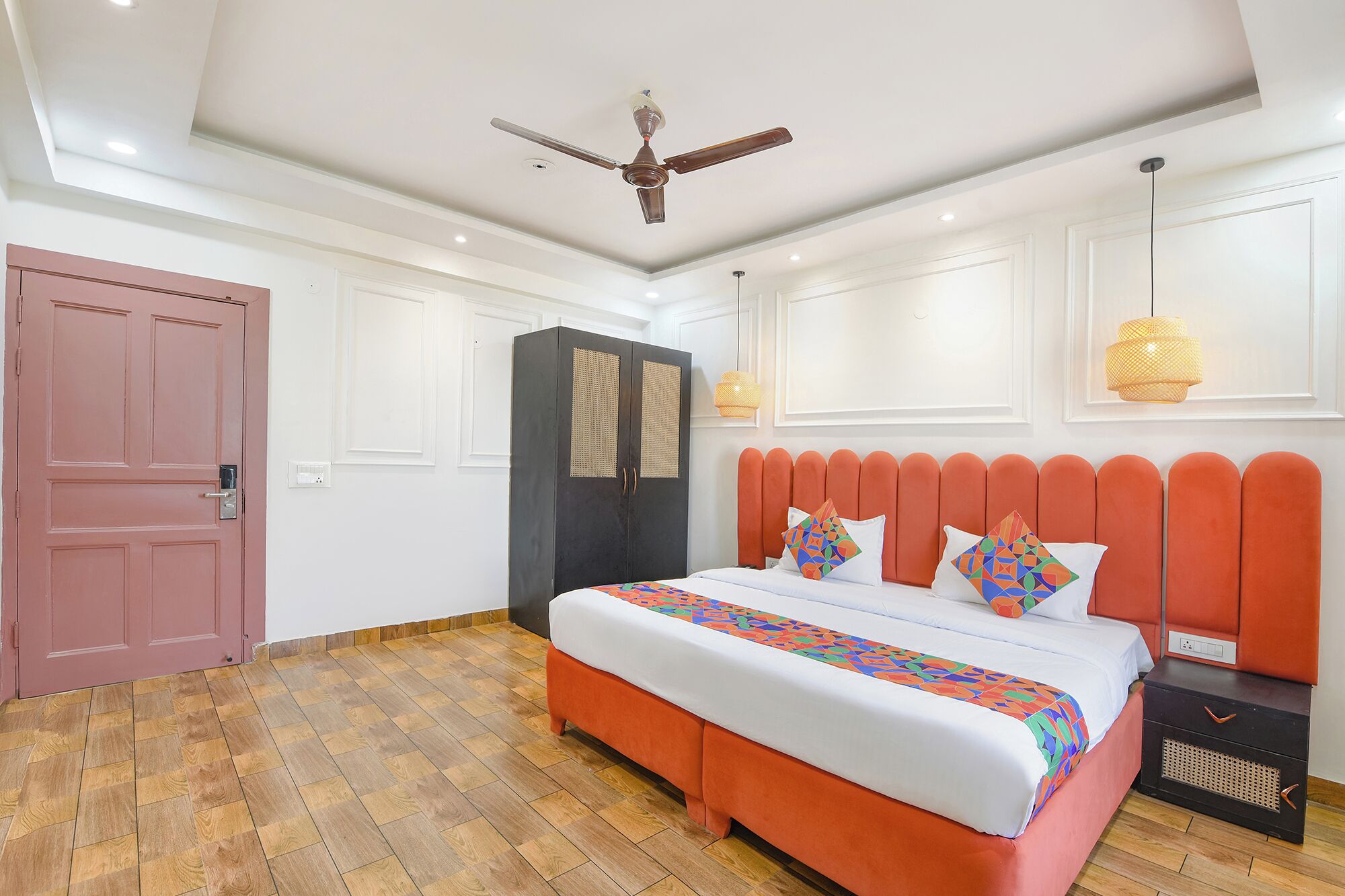 A bedroom with a wooden floor, a bed with orange headboard, a ceiling fan, and a wardrobe.
