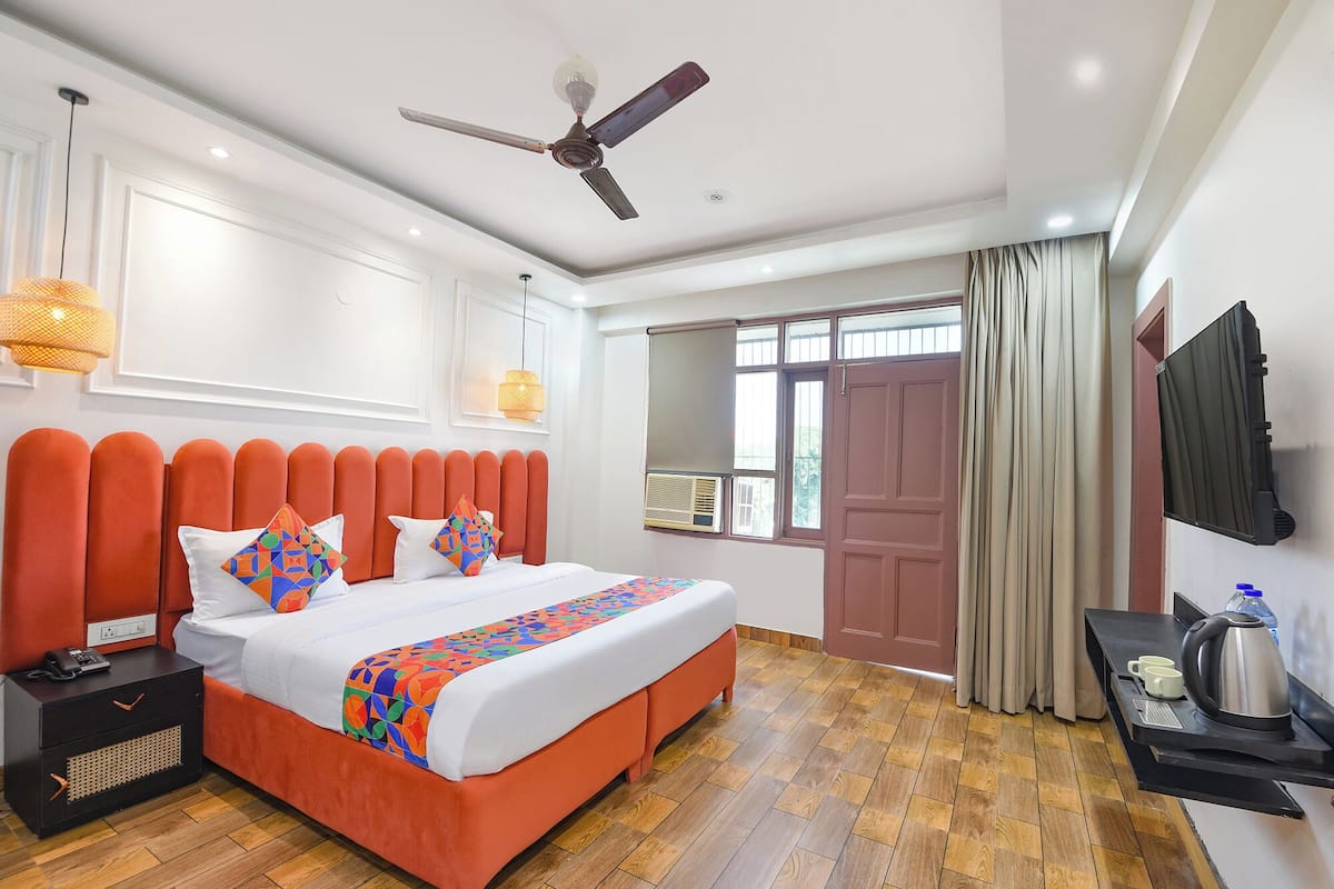 A hotel room with a large bed, a ceiling fan, a window with curtains, a TV, and a small table with a kettle and cups.