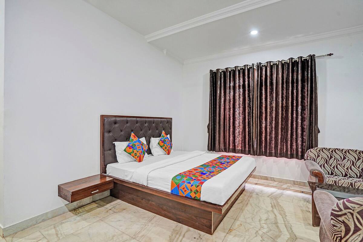 A bedroom with a tufted headboard, a bed with white bedding and colorful geometric pillows, a dark curtain, and a patterned armchair.