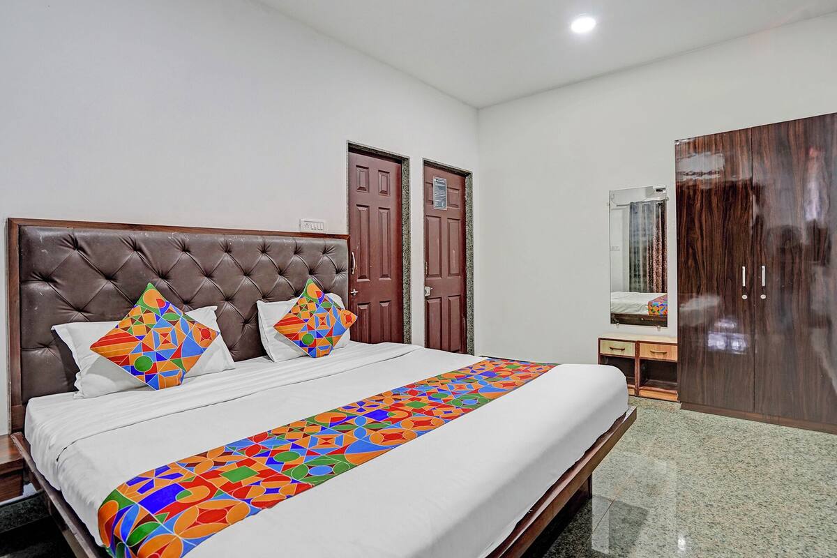 A hotel room with a tufted headboard bed, two doors, a mirror, a wooden wardrobe, and a colorful geometric bed runner.