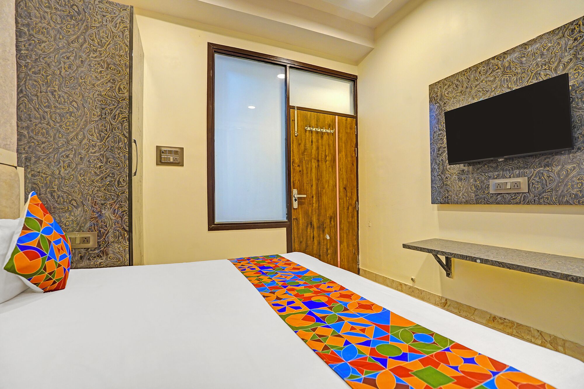 A hotel room with a bed, a TV mounted on the wall, a wooden door, and a patterned wall.
