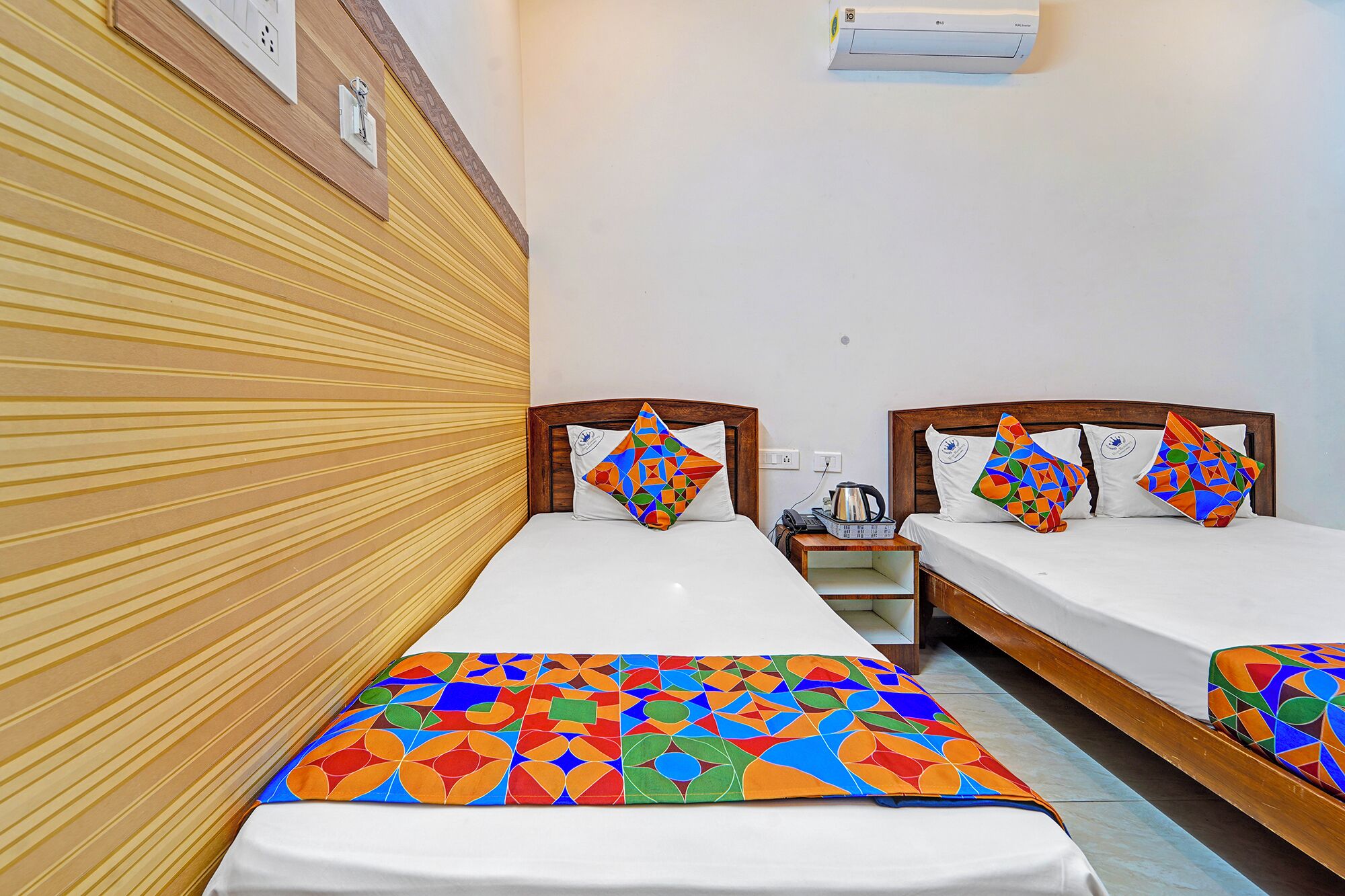 A hotel room with two single beds, wooden headboards, a white wall, and a colorful geometric-patterned bedspread.