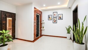 Um corredor amplo com piso de azulejos, porta de madeira, parede de tijolos e vários vasos com plantas.