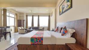Une chambre d’hôtel spacieuse, dotée d’un grand lit, d’une tête de lit en bois, d’un bureau et d’un canapé. Une fenêtre avec des rideaux, un tableau au mur et une vue sur la ville complètent l’ensemble.