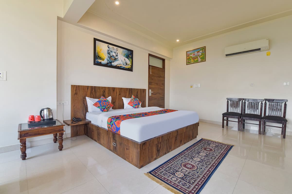 A hotel room with a wooden bed, a nightstand, a desk, two chairs, a patterned rug, and two paintings on the walls.