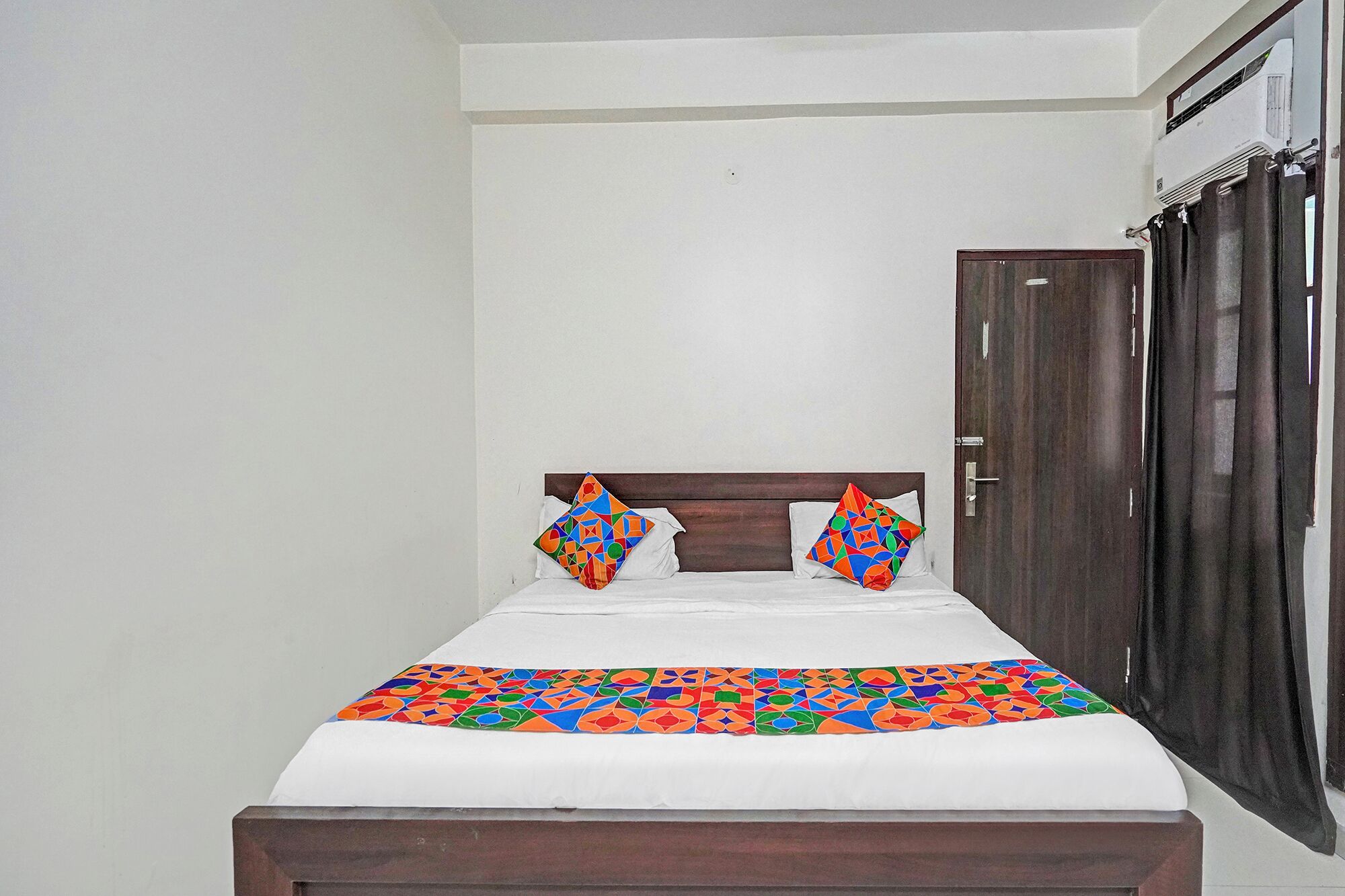 A single bed with a patterned bedspread, two pillows with geometric designs, a wooden headboard, and a dark wooden wardrobe with a door.