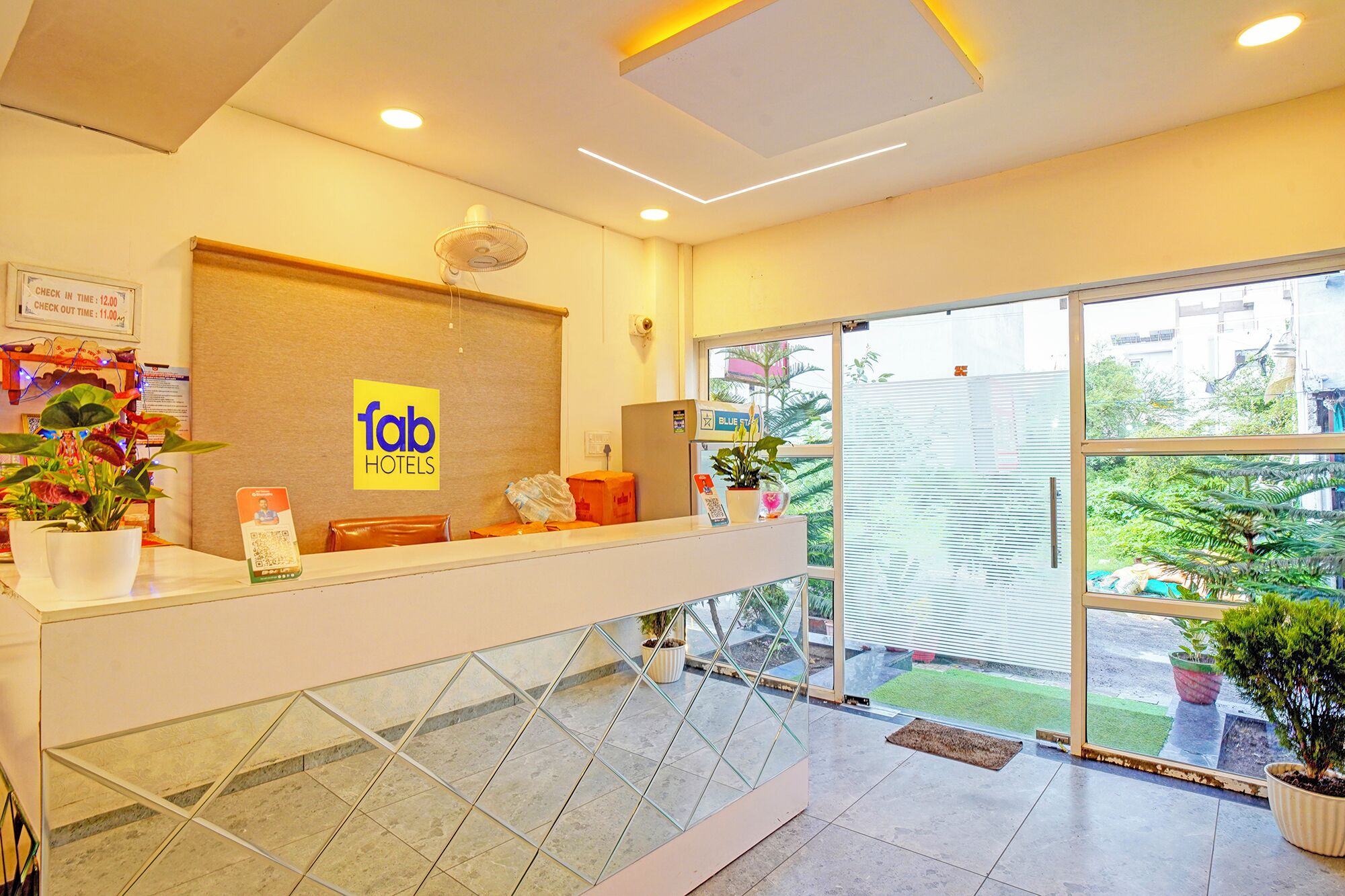 A hotel reception desk with a glass partition, a check-in/out sign, a fan, and potted plants.