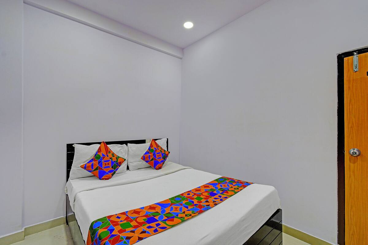 Ein kleines, ordentlich bezogenes Bett mit einer farbenfrohen Bettdecke mit geometrischen Mustern und Kissen, eine weiße Wand und eine Holztür.