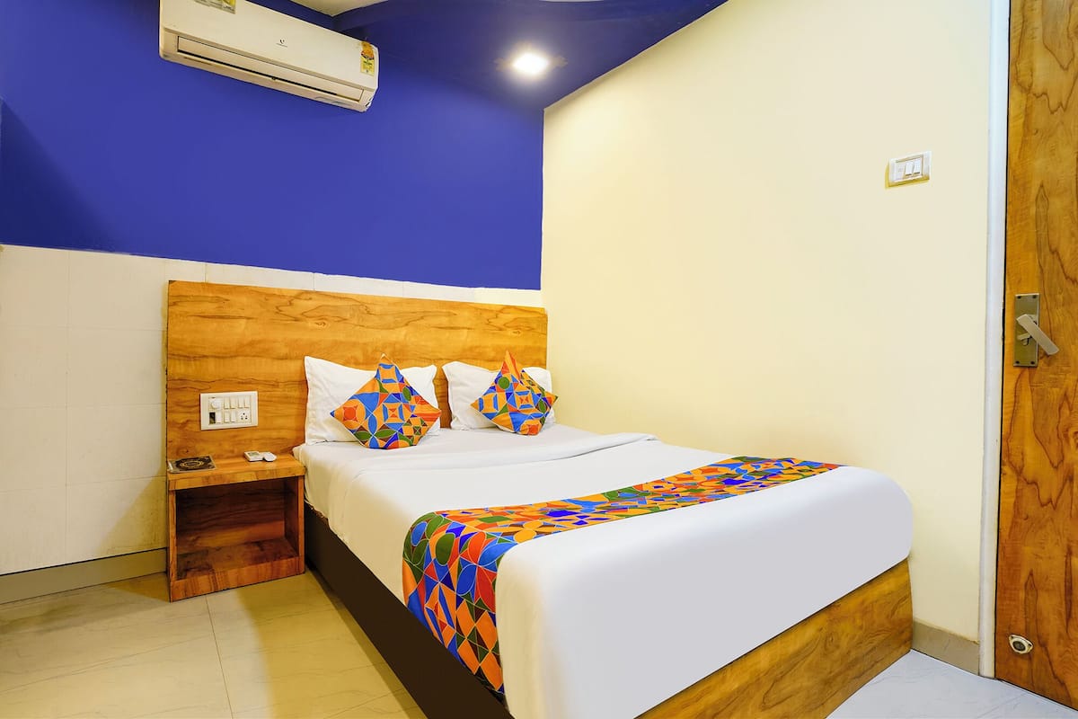 A hotel room with a wooden headboard, a bed with colorful geometric pillows, a bedside table with a phone and a book, and an air conditioning unit on the wall.