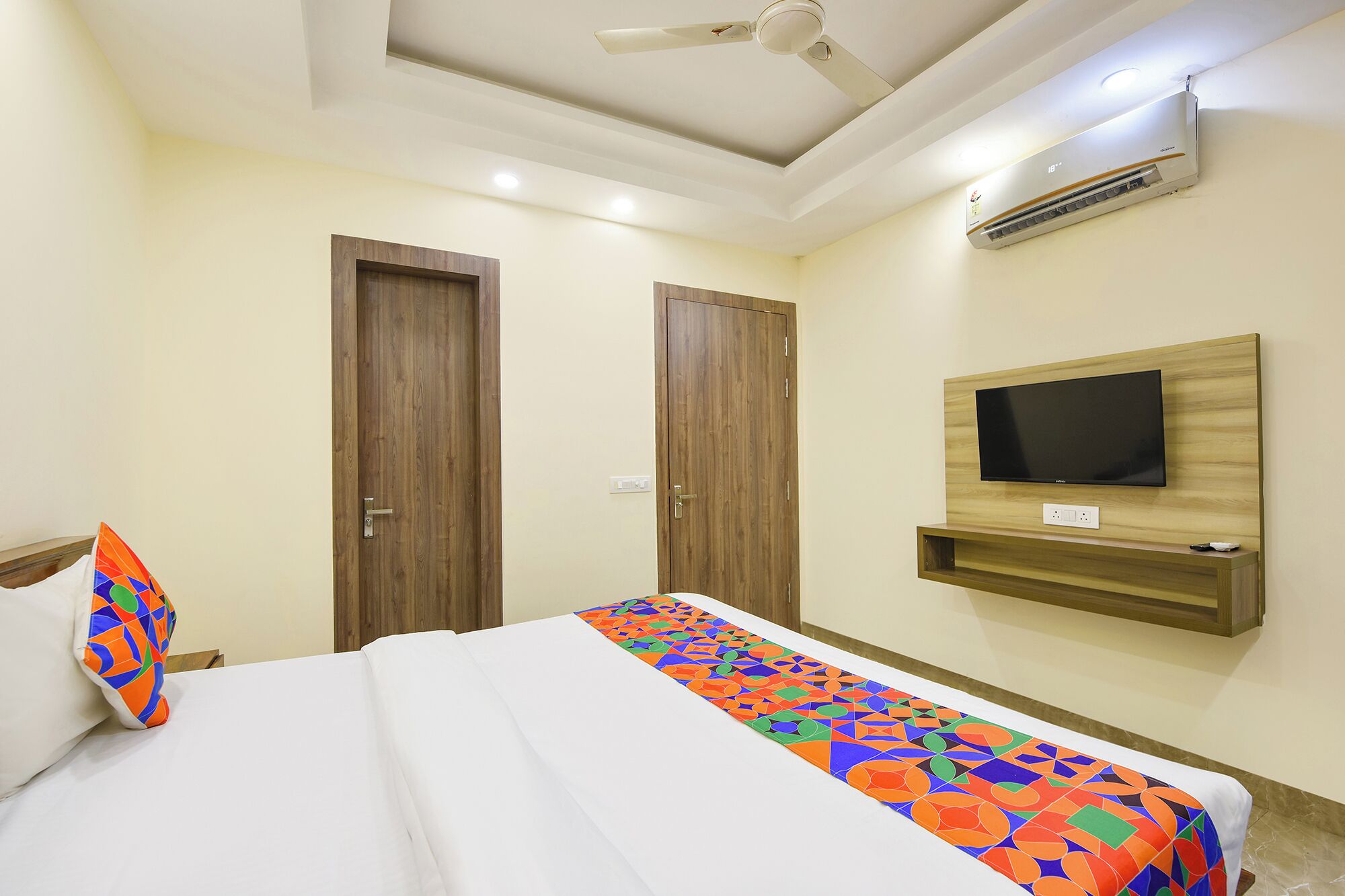 Una habitación de hotel con una cama, un ventilador de techo, un equipo de aire acondicionado, un televisor montado en la pared y una puerta de madera.