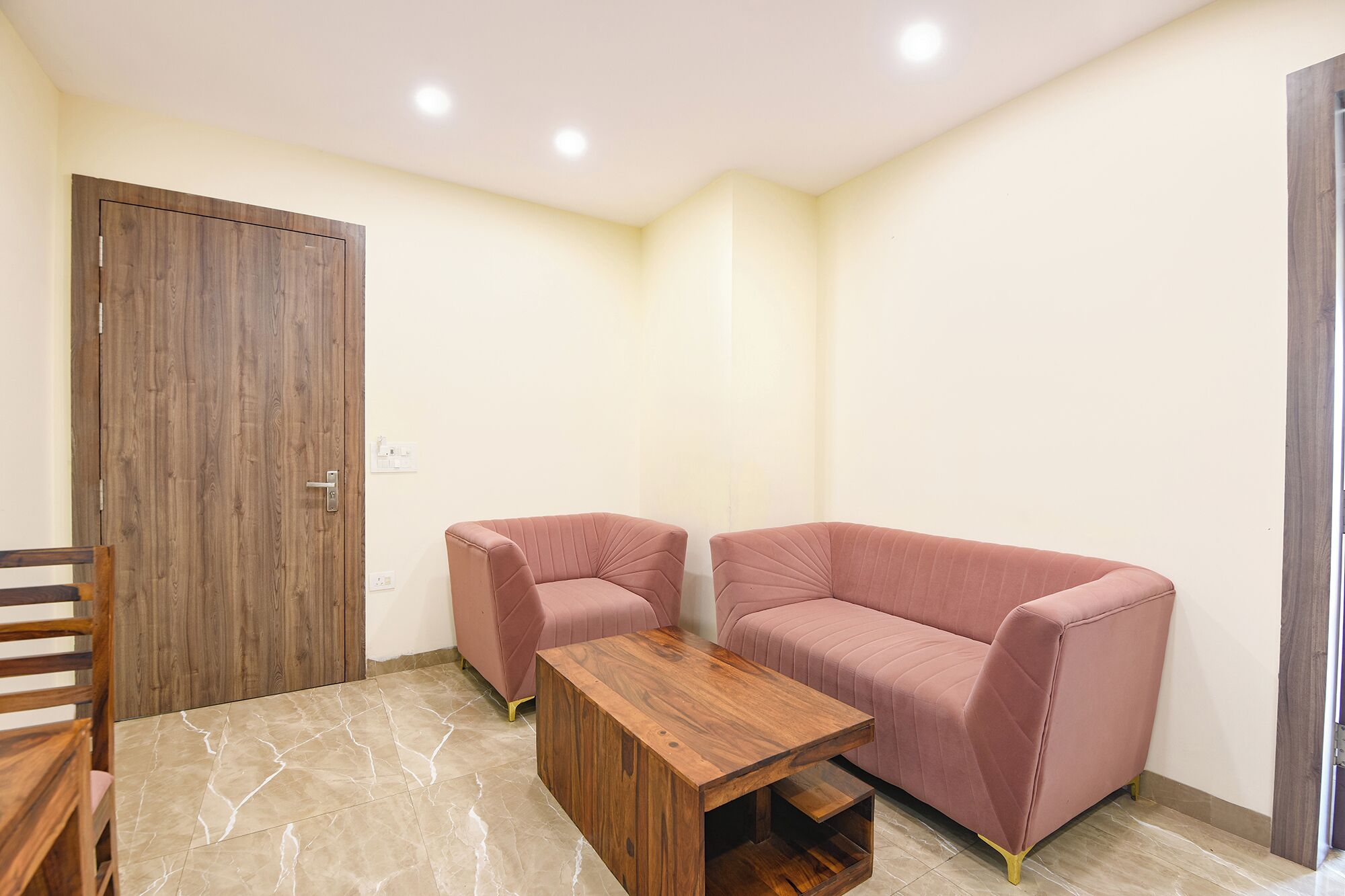 Una stanza piccola e moderna con una porta in legno, un divano rosa, un tavolino in legno e un pavimento piastrellato.