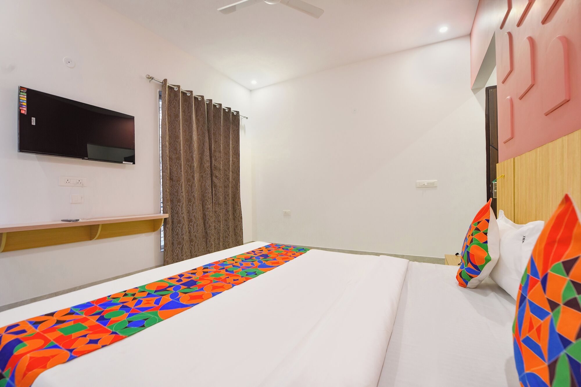 Une chambre à coucher avec un lit, un téléviseur fixé au mur, une fenêtre avec des rideaux et un ventilateur de plafond.