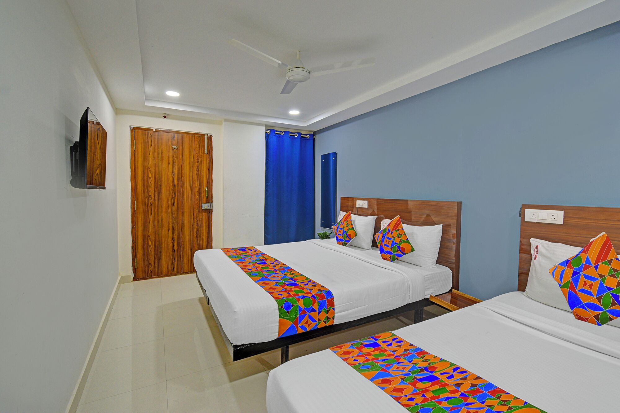 A hotel room with two beds, a ceiling fan, a TV mounted on the wall, and a wooden door.