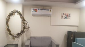 A room with a grey sofa, a small table, a decorative wreath, an air conditioner, and a refrigerator. - Peaceful Escape (Zirakpur)