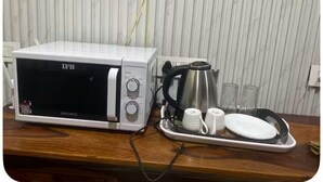 A microwave oven with a tray of white cups and a stainless steel pot on a wooden countertop. - Peaceful Escape (Zirakpur)