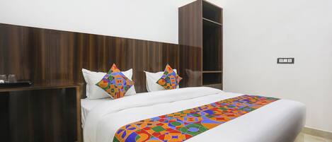 A hotel room with a wooden headboard, a bed with white bedding and colorful geometric pillows, a small painting on the wall, and a wardrobe.