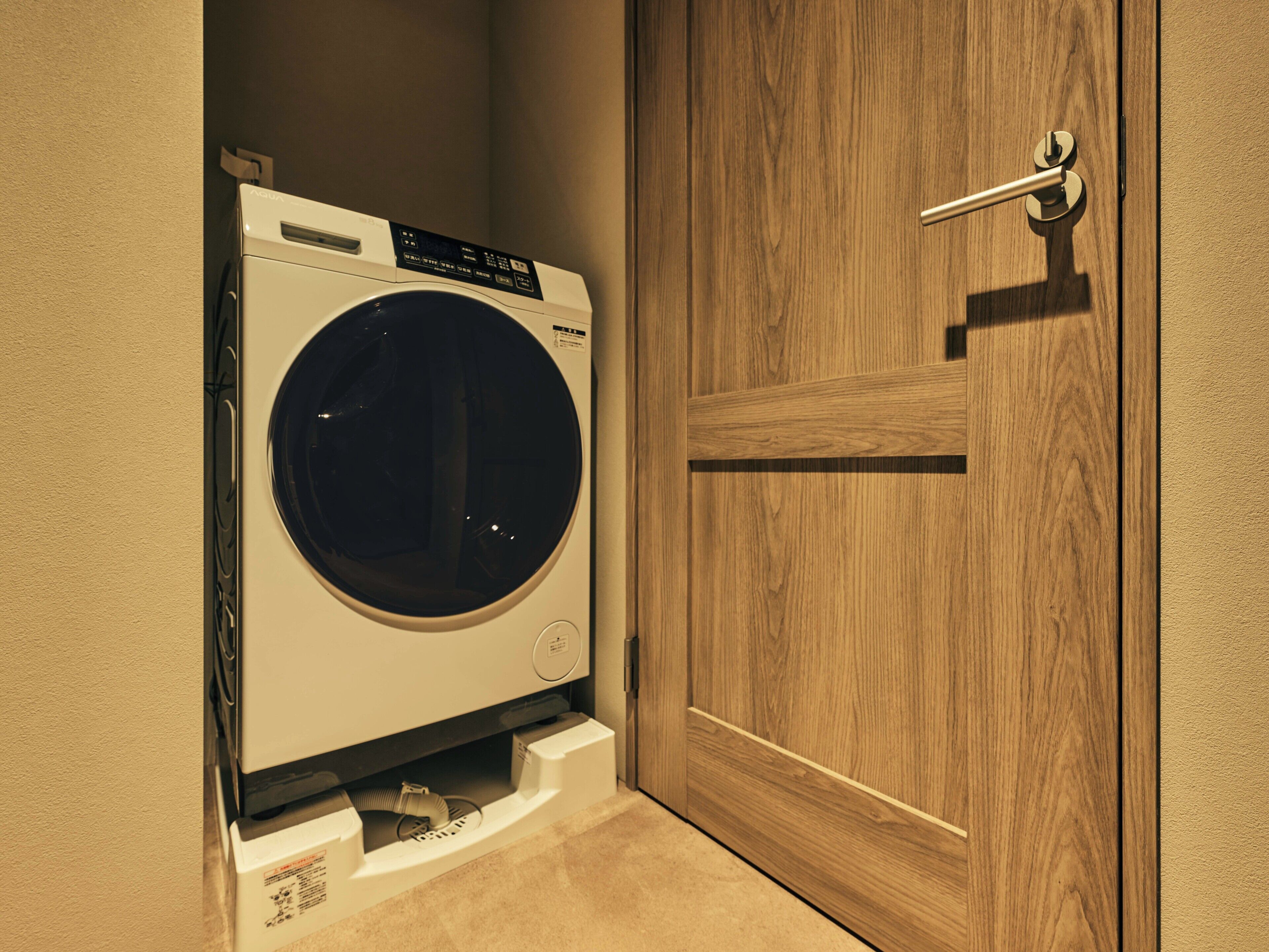 Un lave-linge à chargement frontal installé dans un coin, avec une porte en bois à proximité.