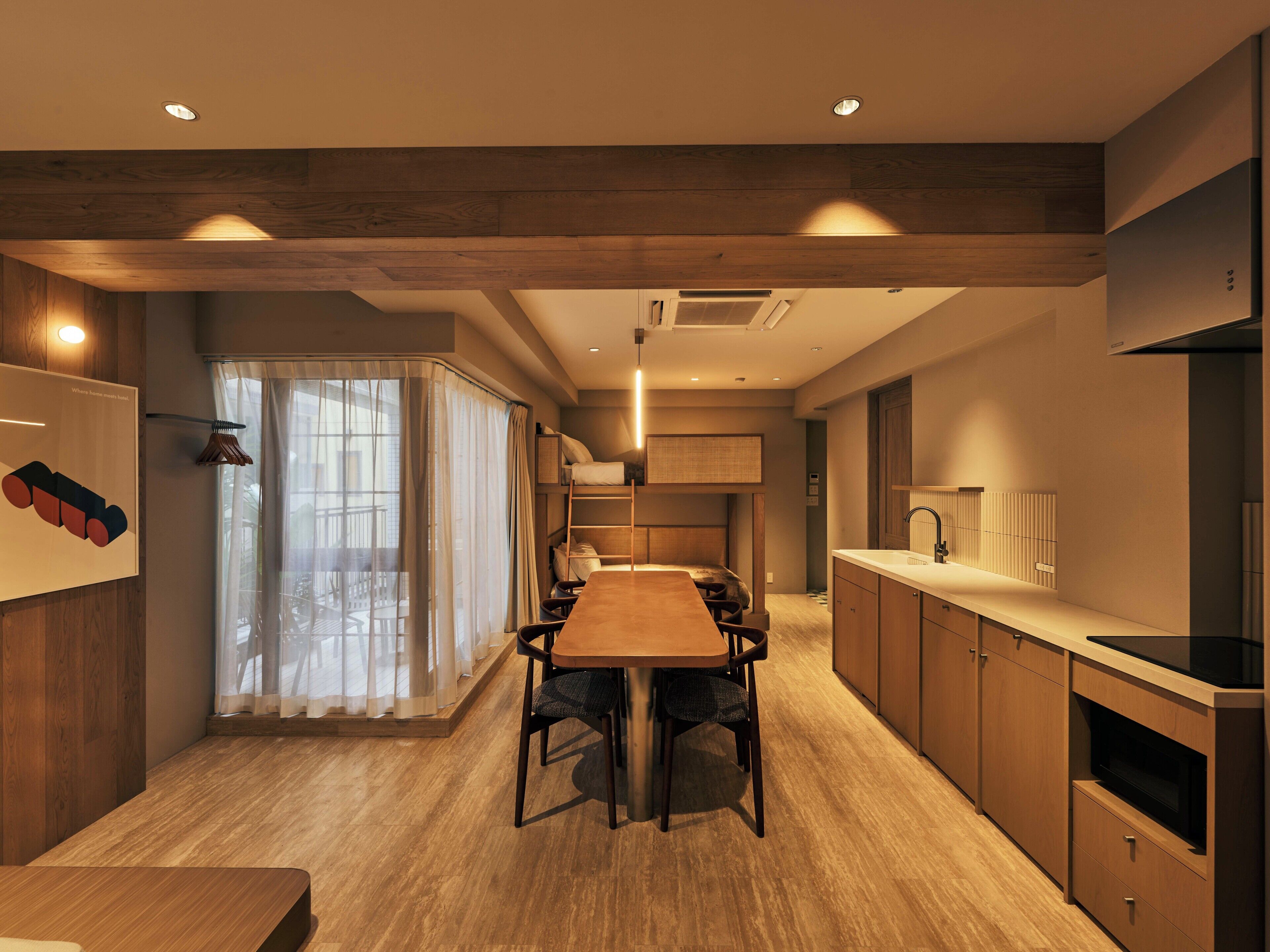 A modern kitchen with wooden cabinets, a dining area with a wooden table and chairs, and a view of a balcony with sheer curtains.