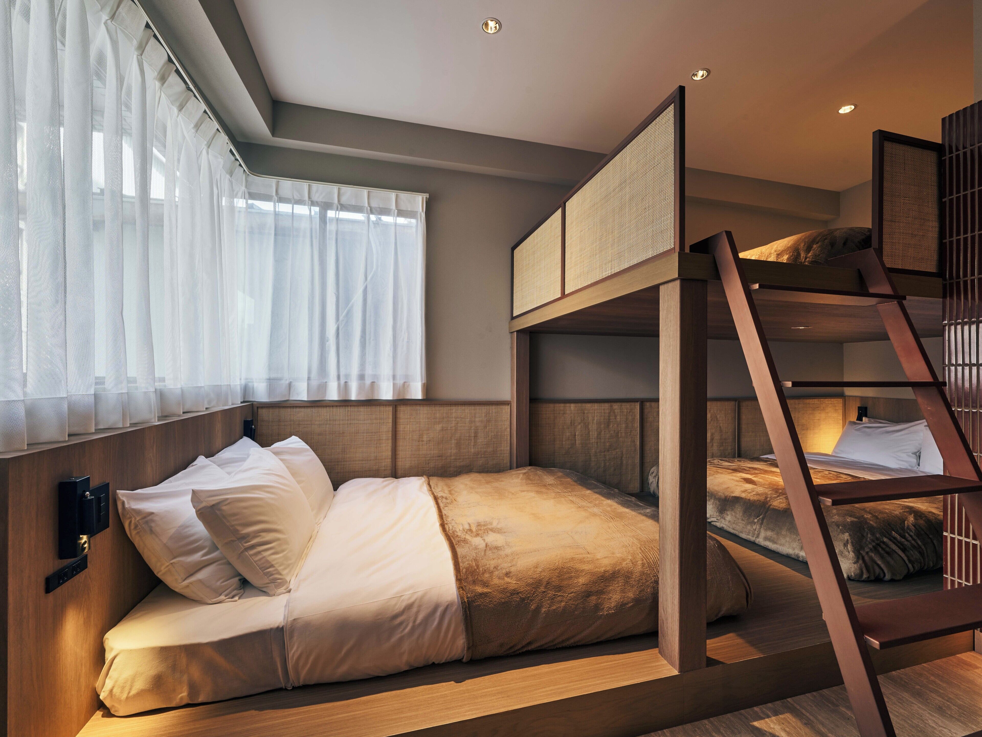A bunk bed room with a wooden ladder, a bedside table, and a window with sheer curtains.
