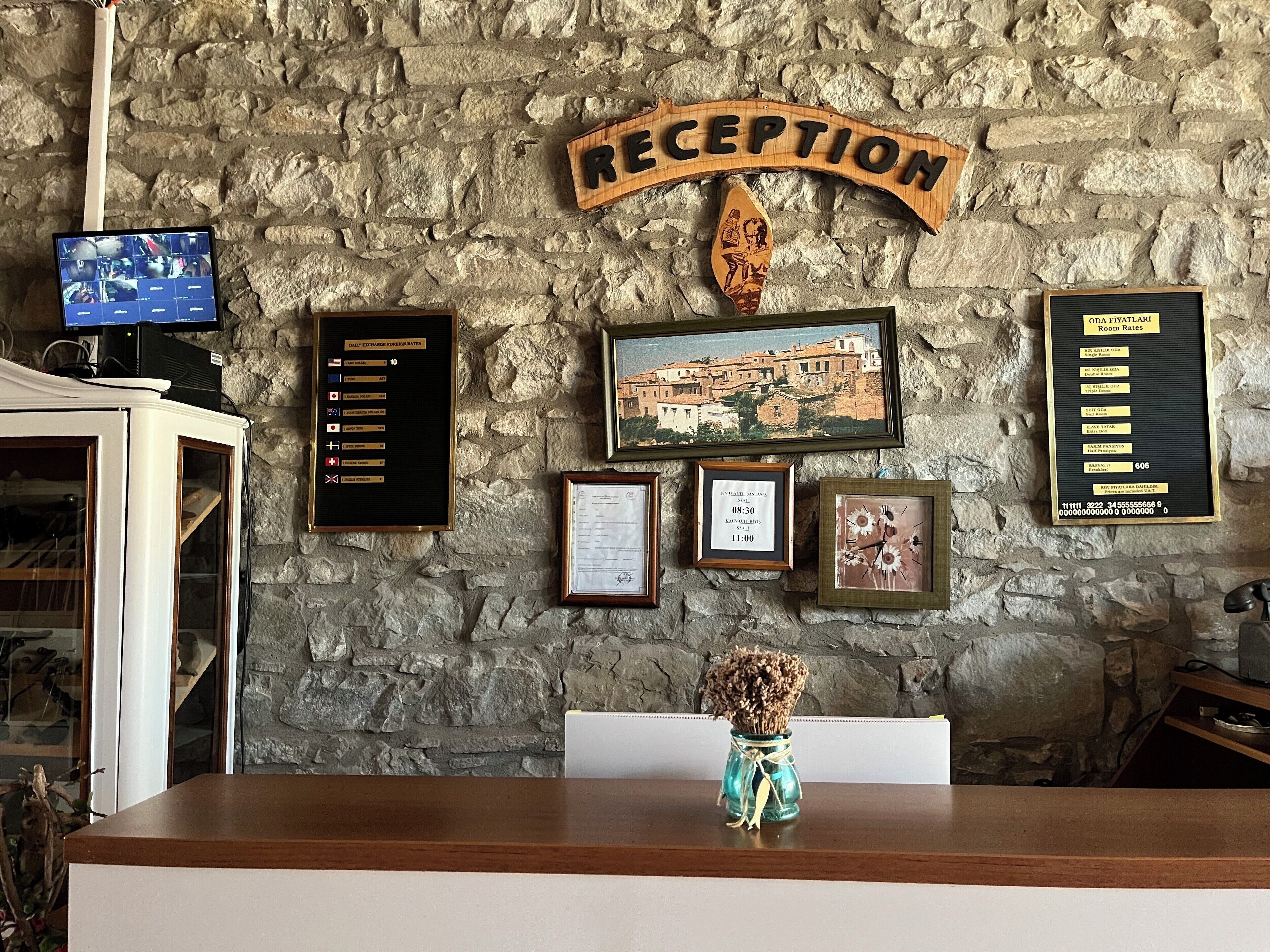 A reception desk with a stone wall background, a framed welcome sign, a framed photograph, a framed certificate, and a framed room rate sign.