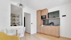 A modern kitchen with wooden cabinets, a microwave, and a coffee maker. There is a white dining table with chairs and a yellow sofa in the living area. - Charming Apartment - 1BR - 2P - Montreuil (Montreuil)