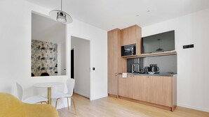 A modern kitchen with wooden cabinets, a microwave, and a coffee maker. There is a dining area with a white table and chairs. - Charming Apartment - 1BR - 2P - Montreuil (Montreuil)