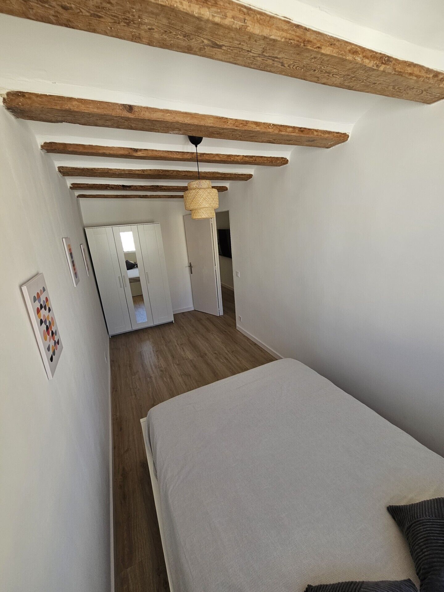 Une chambre à coucher avec un lit, des poutres en bois, un luminaire suspendu et un couloir menant à une salle de bain.
