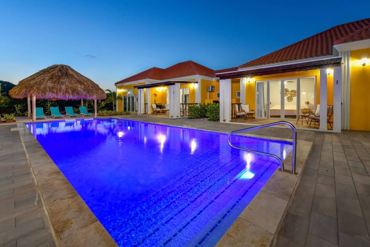A swimming pool with a blue-lit edge, lounge chairs, and a thatched roof structure.