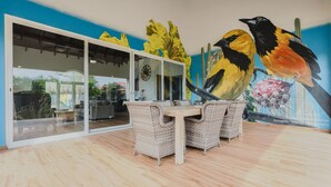 Un espace repas avec un mur en verre, des meubles en osier et une fresque représentant des oiseaux.