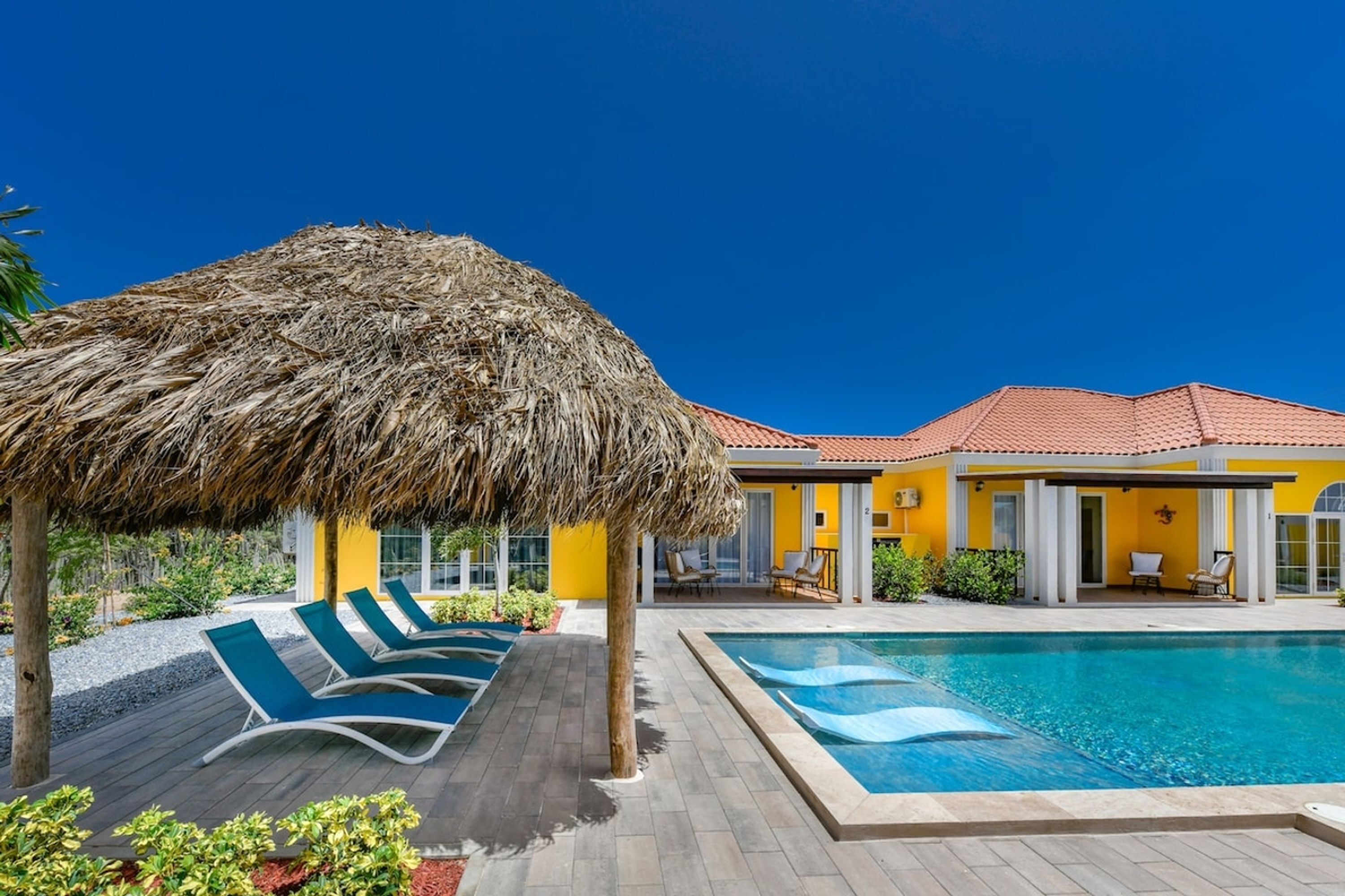 A pool area with lounge chairs, a thatched umbrella, and a yellow building with a red roof.