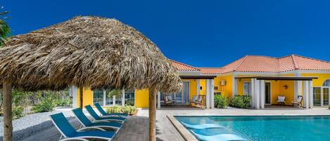 A pool area with lounge chairs, a thatched umbrella, and a yellow building with a red roof.