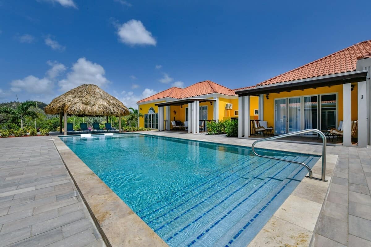 A swimming pool with a thatched roof structure, a covered seating area, and a building with a red-tiled roof.