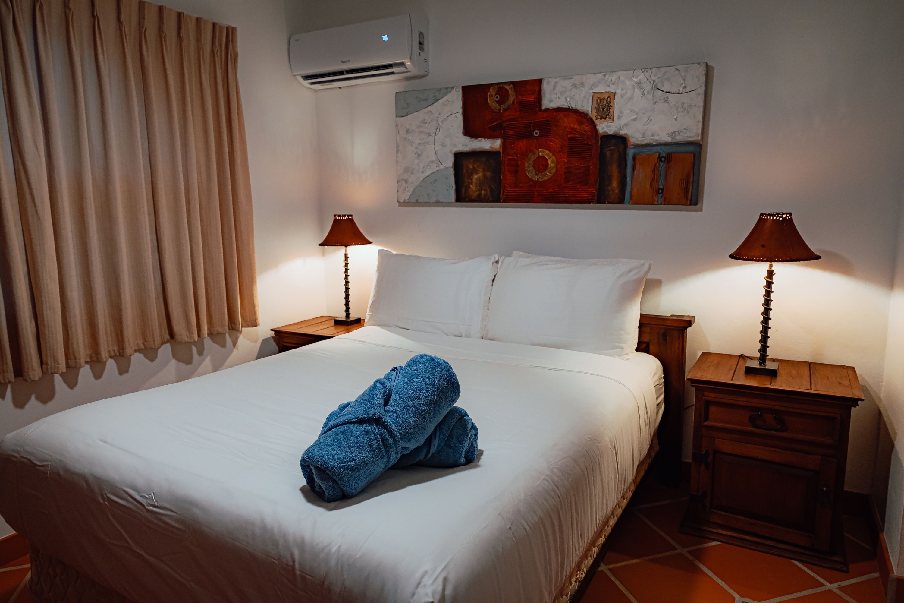 Een hotelkamer met een net opgemaakt bed, twee nachtkastjes met lampen, een wandairconditioner en een schilderij boven het bed.