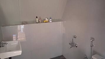 Een kleine, praktische badkamer met een witte wastafel, toilet en douche. Boven de wastafel bevindt zich een plank met toiletartikelen.