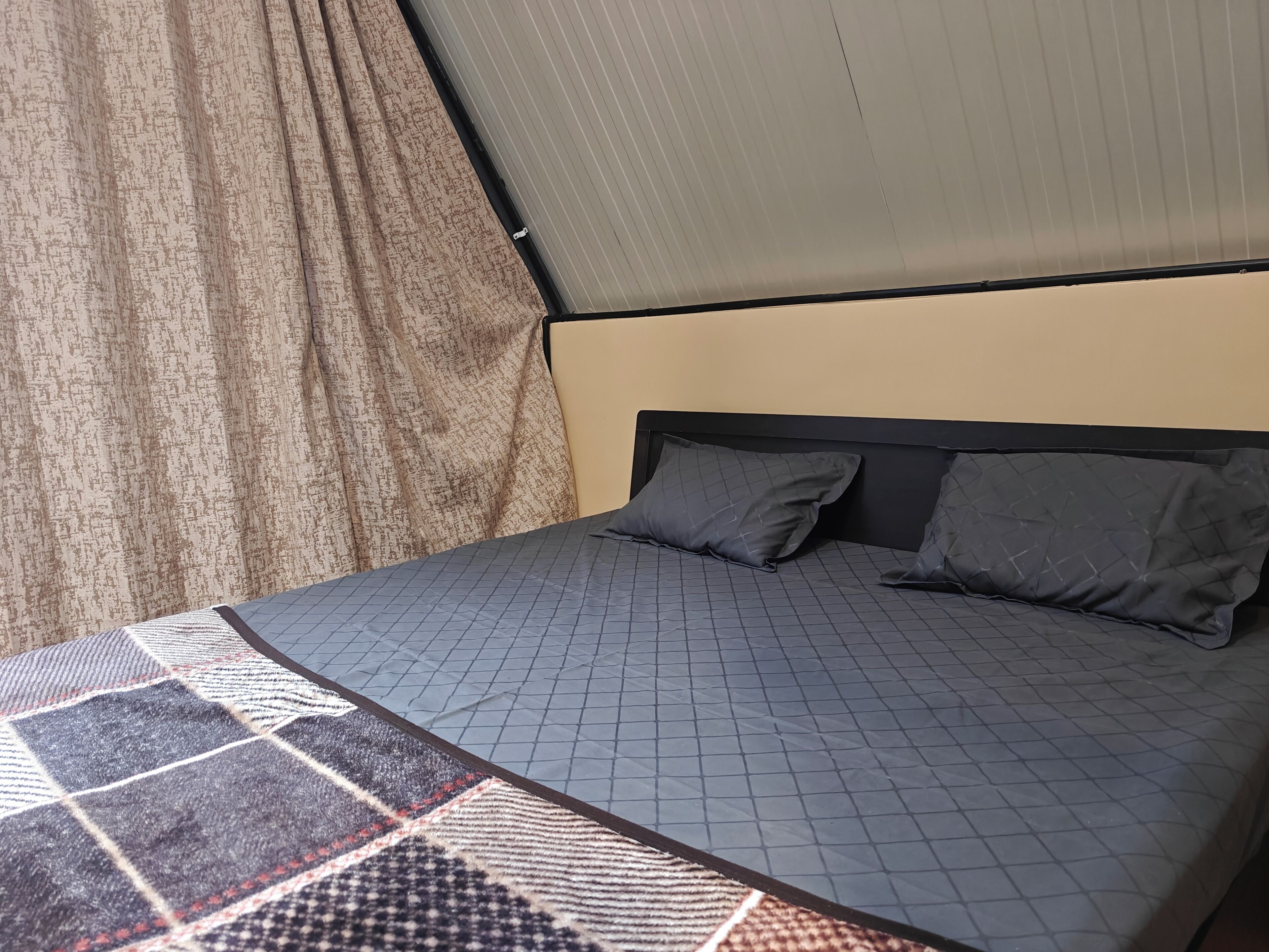 Un letto con copriletto trapuntato grigio e due cuscini, una finestra con tenda e una ventola a soffitto.