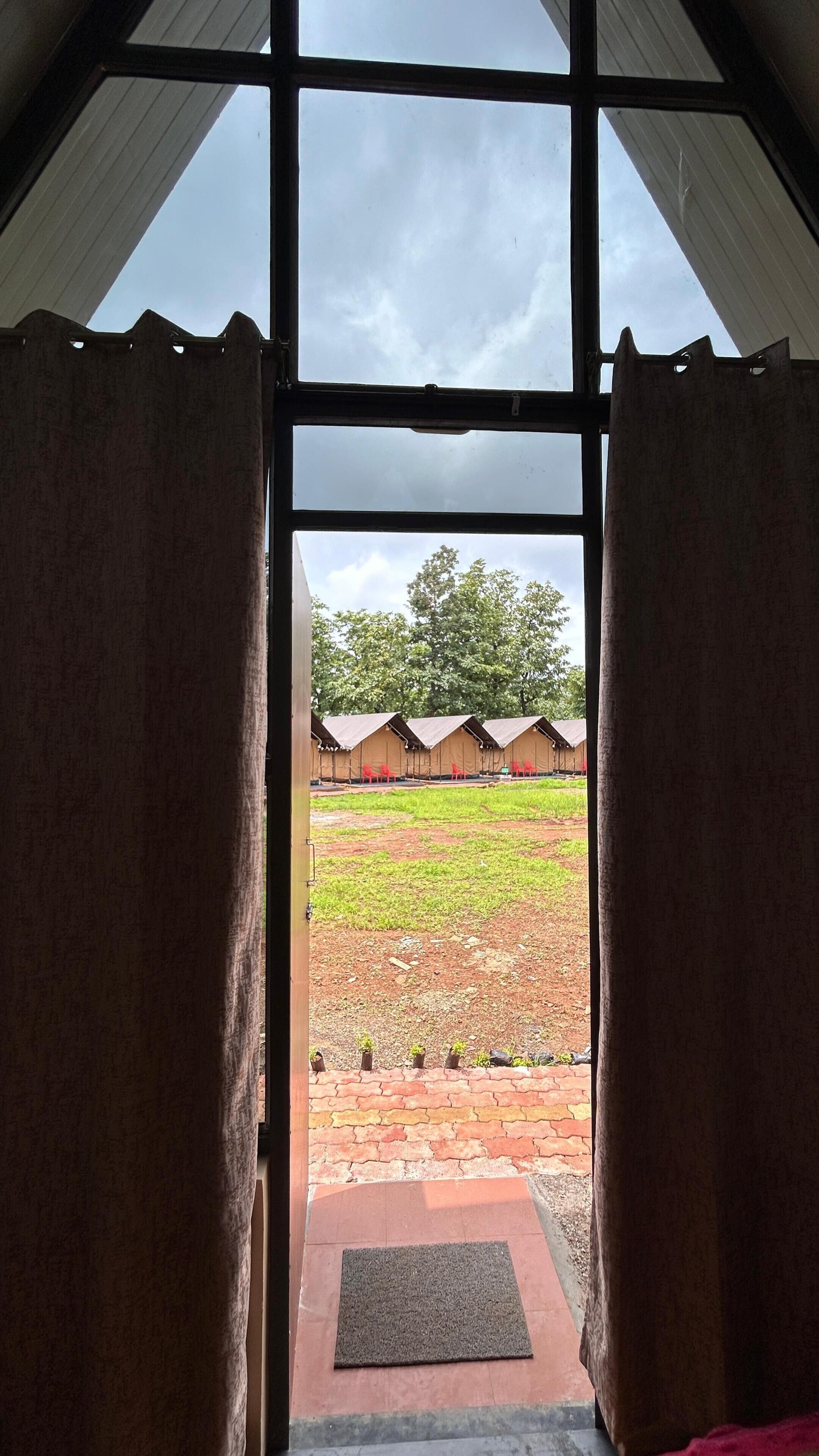 A view through a window with curtains, revealing a grassy area with a brick path and several tents.