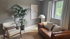 A living room with a brown leather sofa, a wooden armchair, a large potted plant, a floor lamp, and a window with curtains. - Cairn - one bedroom apartment sleeps up to three (Midlothian)
