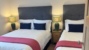 A hotel room with two beds, each with a dark headboard, white bedding, and a pink throw. There are two bedside tables with lamps, and the room has a carpeted floor. - Cairn - one bedroom apartment sleeps up to three (Midlothian)