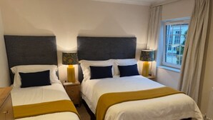 Camera d'albergo con due letti, ognuno con testata scura, biancheria da letto bianca e una coperta color senape. Sono presenti comodini con lampade e una finestra con tende.