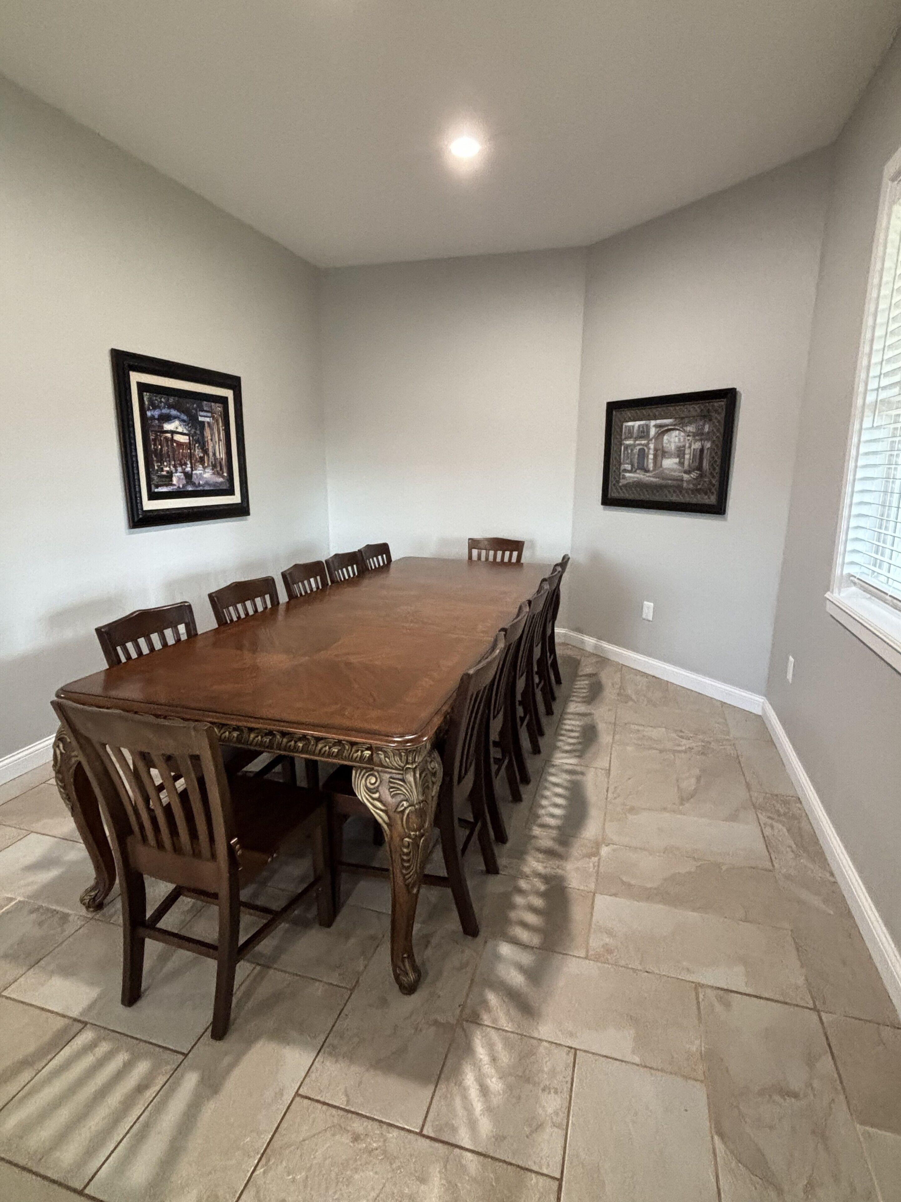 A dining room with a large wooden table surrounded by chairs, two framed pictures on the walls, and a window with blinds.